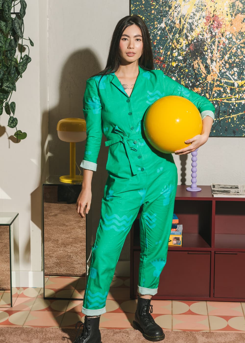 Woman in a green outfit holding a yellow ball in an indoor setting with a colorful abstract painting on the wall.