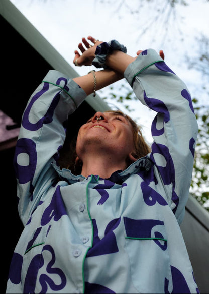 Person wearing a light-coloured shirt with blue patterns, standing outdoors with trees in the background.
