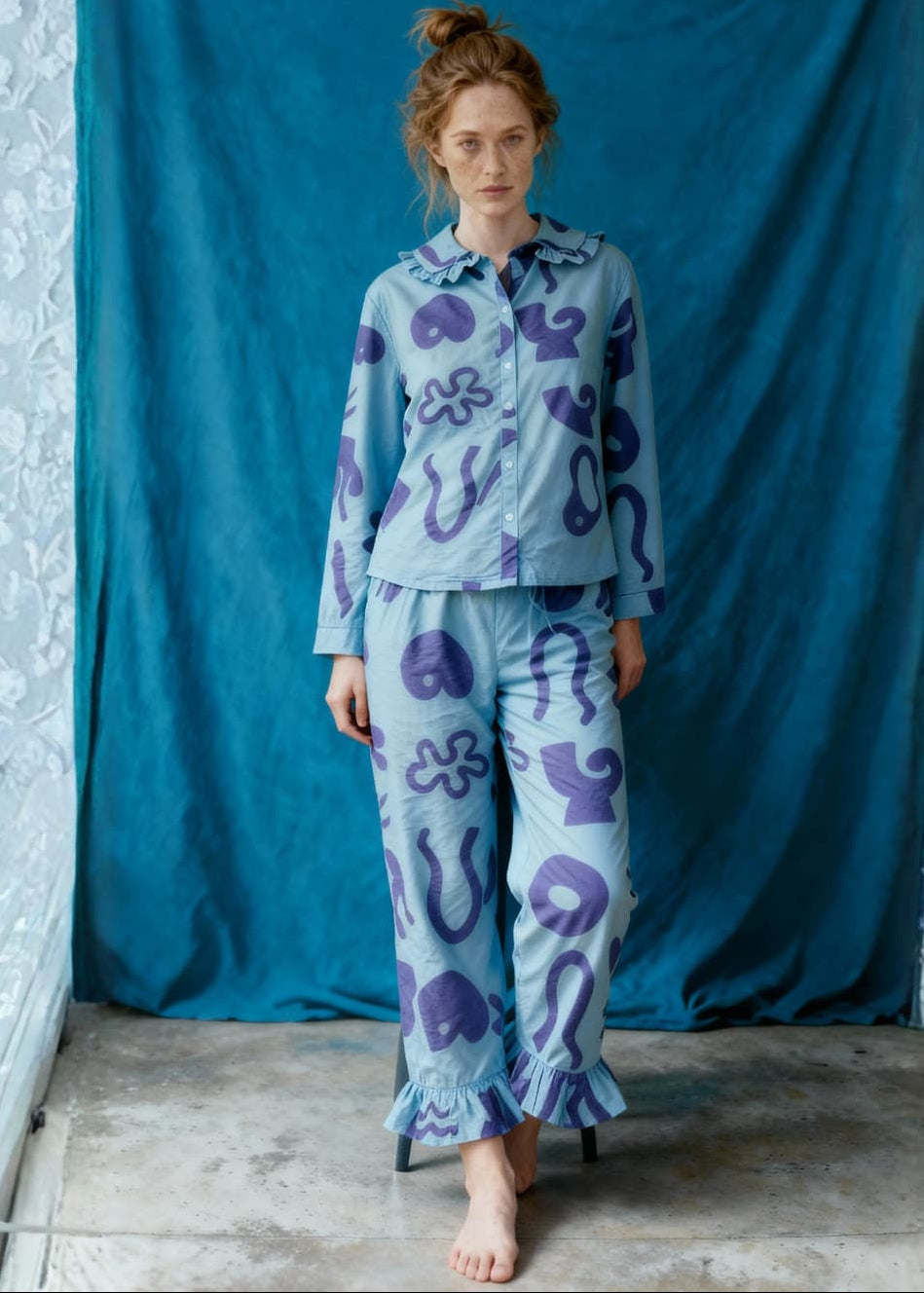 Woman wearing a light blue pyjama set with purple patterns against a blue curtain.
